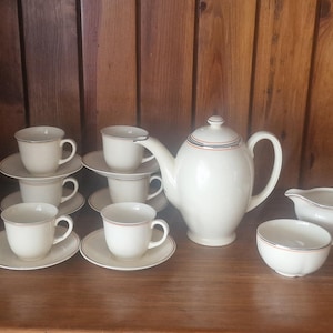May include: A cream tea set comprising a teapot, cups, saucers, a creamer, and a bowl. The teapot and cups feature a thin red and blue stripe. The set is arranged on a wooden surface.