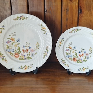 May include: Two white ceramic plates with floral designs. The plates feature a spiral edge and are decorated with colorful flowers in shades of blue, yellow, and orange. The plates are displayed on black metal stands.
