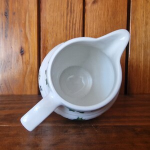 May include: White ceramic creamer with a green and white floral design. The creamer is empty and has a handle.