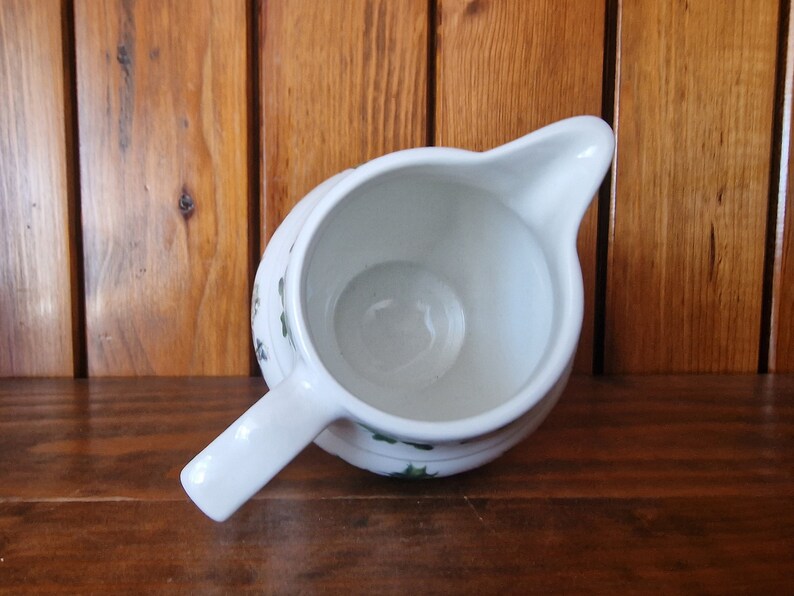 May include: White ceramic creamer with a green and white floral design. The creamer is empty and has a handle.