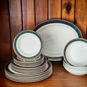 Peut inclure: Une collection de vaisselle en porcelaine blanche avec un motif de bordure bleu et or. L'ensemble comprend des assiettes, des bols et un plat de service de diff&eacute;rentes tailles. Les assiettes sont empil&eacute;es et les bols sont empil&eacute;s.