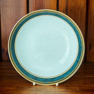 Peut inclure: Une assiette en c&eacute;ramique blanche avec une bordure turquoise et dor&eacute;e. L'assiette est ronde et poss&egrave;de un centre l&eacute;g&egrave;rement creux. La bordure turquoise est orn&eacute;e d'une garniture dor&eacute;e et de petits &eacute;l&eacute;ments d&eacute;coratifs. L'assiette est pr&eacute;sent&eacute;e sur un support blanc.