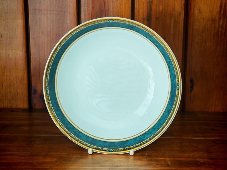 Peut inclure: Une assiette en c&eacute;ramique blanche avec une bordure turquoise et dor&eacute;e. L'assiette est ronde et poss&egrave;de un centre l&eacute;g&egrave;rement creux. La bordure turquoise est orn&eacute;e d'une garniture dor&eacute;e et de petits &eacute;l&eacute;ments d&eacute;coratifs. L'assiette est pr&eacute;sent&eacute;e sur un support blanc.