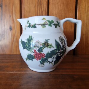 May include: White ceramic pitcher with a green holly and red berry design. The pitcher has a rounded body and a handle.