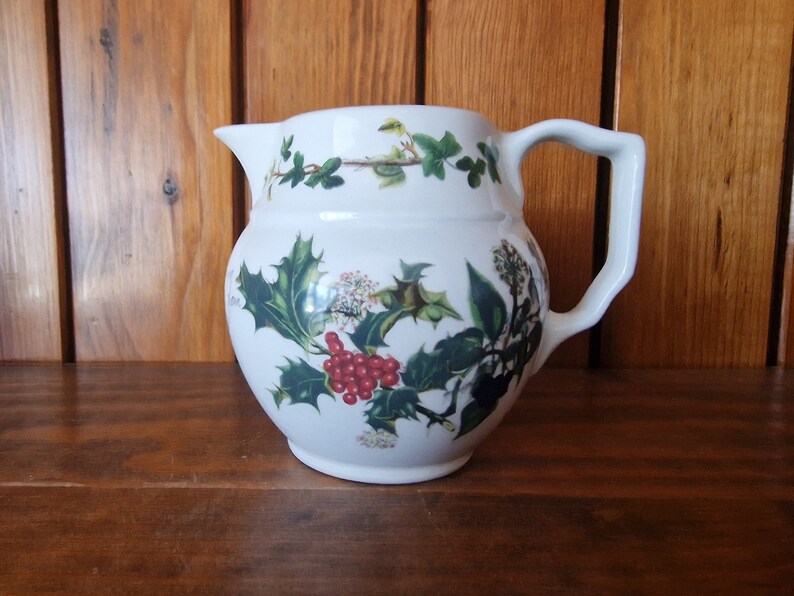 May include: White ceramic pitcher with a green holly and red berry design. The pitcher has a rounded body and a handle.