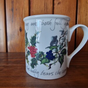 May include: White ceramic mug with a Christmas-themed design. The mug features a holly sprig with red berries, a sprig of ivy with blue berries, and the text "They are both full grown, the holly bears the crown".