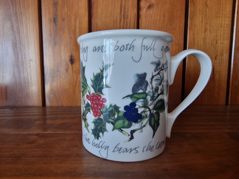 May include: White ceramic mug with a Christmas-themed design. The mug features a holly sprig with red berries, a sprig of ivy with blue berries, and the text "They are both full grown, the holly bears the crown".