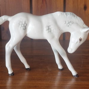 May include: A white ceramic foal figurine with grey floral detailing. The foal is depicted in a standing pose, with its head lowered. The hooves and ears are black. This figurine is a decorative item.