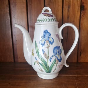 Könnte beinhalten: Weiße Keramik-Teekanne mit Deckel, verziert mit blauen Irisblüten und grünen Blättern. Die Teekanne hat einen gebogenen Ausguss und Henkel, mit einem Schmetterlingsdesign auf dem Deckel. Der Sockel hat einen kleinen Sockel.