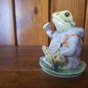 May include: A ceramic figurine of a frog wearing a purple coat and holding a small white object. The frog is sitting on a green lily pad.