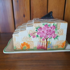 May include: A ceramic cheese dish shaped like a brick wall with a sundial on top. The wall is decorated with flowers and a tree with pink blossoms. The dish has a matching plate.