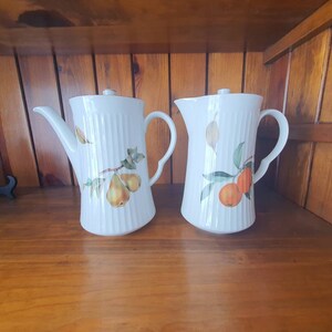May include: Two white ceramic coffee pots with handles. One pot features a pear design and the other an orange design. Both pots have a ribbed texture.