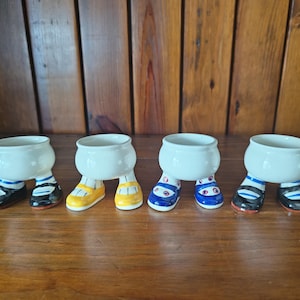 Peut inclure: Quatre petits bols en céramique blanche avec des pieds peints avec différents motifs de chaussures. Les chaussures comprennent des accents noirs et blancs, jaunes, bleus et rouges. Les bols sont disposés sur une surface en bois.