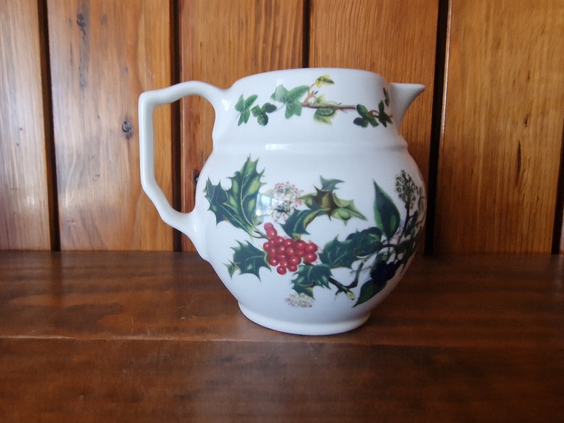 May include: White ceramic pitcher with a floral design featuring holly, berries, and ivy. The pitcher has a rounded body and a handle.