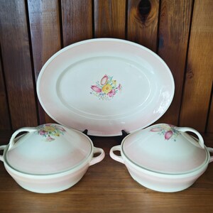 May include: A ceramic serving set consisting of one large oval platter and two matching lidded bowls. Each piece features a cream-coloured base with a soft pink gradient rim and a central floral motif of yellow, pink, and green flowers.