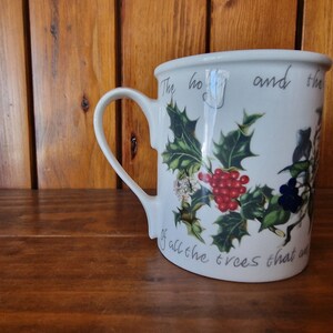 May include: White ceramic mug with a green holly and red berry design. The mug has a handle and reads "The holly and the ivy of all the trees that are".