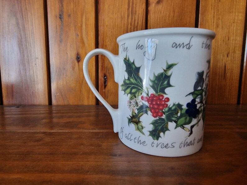 May include: White ceramic mug with a green holly and red berry design. The mug has a handle and reads "The holly and the ivy of all the trees that are".