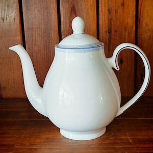May include: White ceramic teapot with a rounded body, curved spout, and a handle with a black trim. The lid has a small knob and a blue and white geometric design. The teapot sits on a wooden surface.