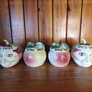 May include: Set of four ceramic apple-shaped containers with painted faces and lids. The lids are labeled "Apple Sauce" and "Pie Sauce".