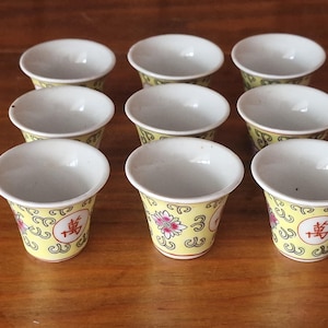 May include: Set of nine small white ceramic teacups with yellow and pink floral designs and Chinese characters. The cups have a rounded bottom and flared rim.