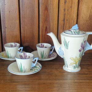 May include: A vintage tea set comprising a cream-coloured teapot and three teacups with saucers. The set is decorated with purple, yellow, and green floral designs, and light blue accents. The teapot has a unique angular handle.