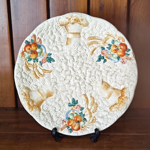 May include: Cream ceramic plate with embossed wheat and fruit designs. The plate has a textured surface with detailed relief patterns. Decorative elements include wheat sheaves and arrangements of oranges and other fruits, accented with blue and green details.