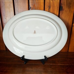 Peut inclure: Un grand plat de service ovale blanc avec un bord l&eacute;g&egrave;rement sur&eacute;lev&eacute;. Le dessous du plat est visible, montrant une indentation ovale centrale et un petit embl&egrave;me dor&eacute;. Le plat est pr&eacute;sent&eacute; sur un support noir sur fond de bois.
