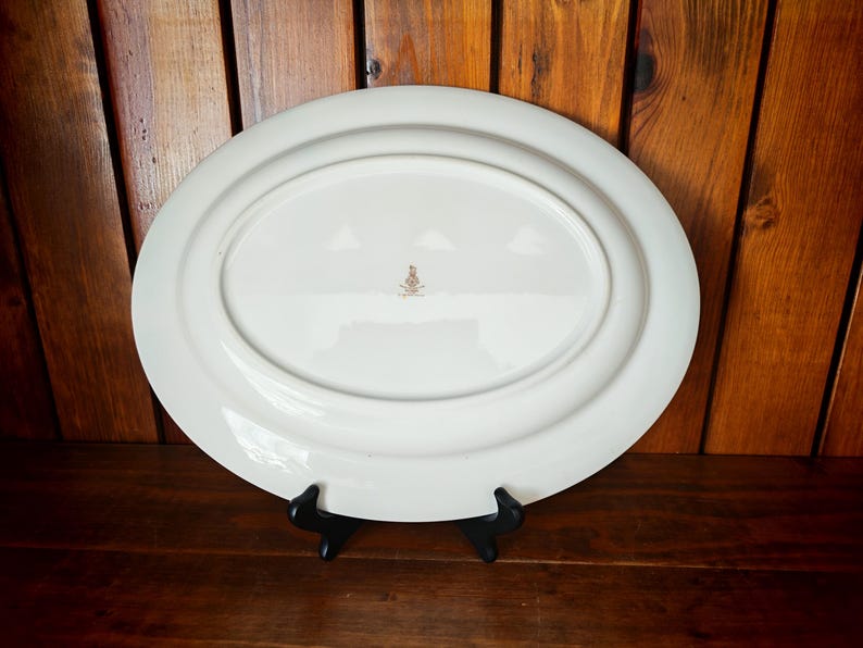 Peut inclure: Un grand plat de service ovale blanc avec un bord l&eacute;g&egrave;rement sur&eacute;lev&eacute;. Le dessous du plat est visible, montrant une indentation ovale centrale et un petit embl&egrave;me dor&eacute;. Le plat est pr&eacute;sent&eacute; sur un support noir sur fond de bois.