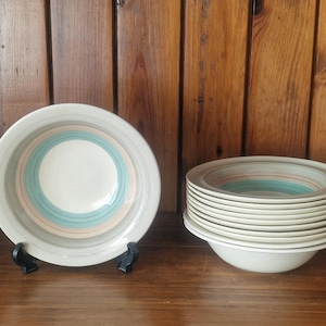 May include: A collection of cream-coloured ceramic bowls featuring concentric rings of grey, teal, and peach. One bowl is displayed on a black stand, and a stack of bowls is next to it.