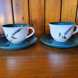 May include: Two white teacups with a green and blue floral design on a teal saucer. The teacups have a white background with a green and blue floral design.