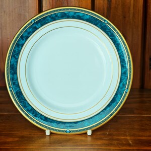 Peut inclure: Une assiette en c&eacute;ramique blanche avec une bordure turquoise et dor&eacute;e. L'assiette pr&eacute;sente un motif marbr&eacute; turquoise avec une bordure dor&eacute;e et de petits accents turquoise. L'assiette est pr&eacute;sent&eacute;e sur une surface en bois.