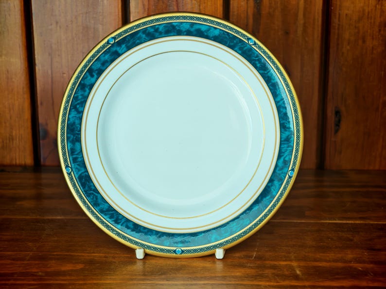 Peut inclure: Une assiette en c&eacute;ramique blanche avec une bordure turquoise et dor&eacute;e. L'assiette pr&eacute;sente un motif marbr&eacute; turquoise avec une bordure dor&eacute;e et de petits accents turquoise. L'assiette est pr&eacute;sent&eacute;e sur une surface en bois.