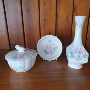 May include: A set of three white ceramic pieces with floral designs. The pieces include a small bowl with a lid, a small plate, and a vase. All three pieces feature a floral design with pink, blue, and green flowers.