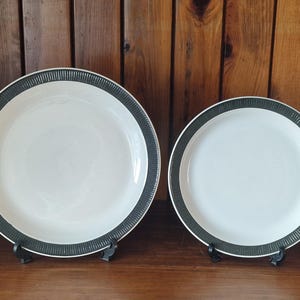 Peut inclure: Deux assiettes en céramique blanche avec une bordure texturée vert foncé. Les assiettes sont présentées sur des supports noirs sur un fond en bois. L'une des assiettes est plus grande que l'autre.
