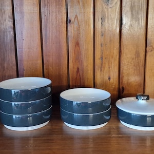 May include: A collection of three ceramic serving bowls and a lidded dish. The bowls are dark grey with white rims and interiors. The lid has a black knob. The items are on a wooden surface against a wooden backdrop.