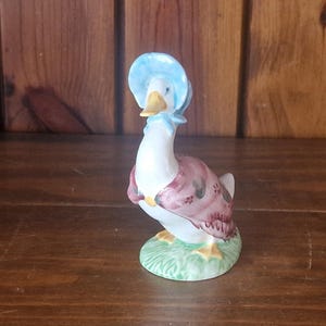 以下が含まれることがあります： 水色の帽子とピンクと紫の模様のショールを着たガチョウの陶器製置物。ガチョウは、黄色のくちばしと足で、緑の草の台座の上に立っています。置物は木製の背景に置かれています。