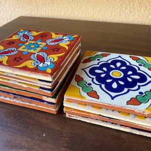 May include: Stacks of square, hand-painted ceramic tiles with colorful floral designs. One stack features a red background with blue, yellow, and white accents. The other stack has a white background with blue and green floral patterns. These tiles are perfect for home decor.