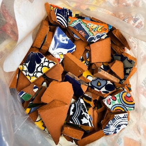 Puede incluir: Una bolsa de plástico transparente llena de trozos rotos de baldosas de cerámica decorativas. Las baldosas presentan varios diseños en azul, amarillo, naranja y blanco. Los trozos rotos se mezclan con fragmentos de terracota.