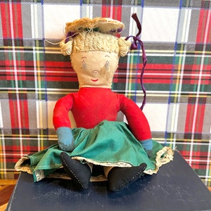 Puede incluir: Una muñeca vintage hecha a mano con pelo de lana rubio, un vestido rojo, una falda verde con ribete de encaje y zapatos de tela azul. La muñeca está sentada sobre una superficie azul.