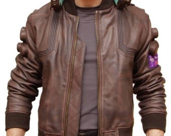 May include: Brown leather bomber jacket with a quilted lining and a ribbed collar and cuffs. The jacket features a zip-up front, a high collar with a green lining, and decorative details on the sleeves. A purple patch with a bird is on the upper arm.