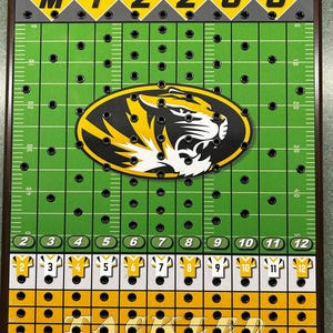 May include: A green and yellow football game board with a black and white tiger mascot in the center. The board has a grid of holes for pegs and the words "MIZZOU" and "TACKLED" printed on it.