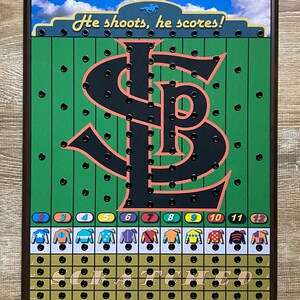 May include: A wooden game board with a green field and a black and red "S" shaped track. The board has a title that reads "He shoots, he scores!" and a grid with numbers 2 through 12. There are 12 colorful jockey silks with numbers 2 through 12. The board has a brown frame.