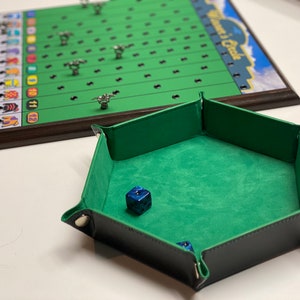 May include: A green felt-lined dice tray with a blue dice inside. The tray is hexagonal and has black leather edges with silver metal snaps.