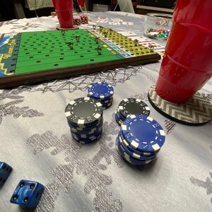 May include: A game board with a green field and white lines, surrounded by a brown wooden frame. The board is titled "Winners & Sinners". There are blue and black poker chips stacked on a white tablecloth.