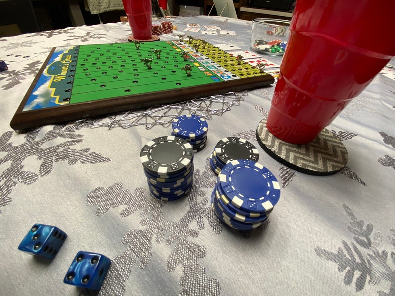 May include: A game board with a green field and white lines, surrounded by a brown wooden frame. The board is titled "Winners & Sinners". There are blue and black poker chips stacked on a white tablecloth.