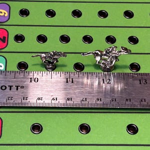 May include: Two silver horse and jockey game pieces on a green game board with black holes. A ruler is placed below the pieces, showing the pieces are about 1 inch tall.