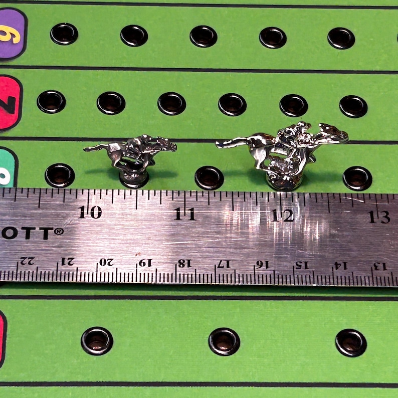 May include: Two silver horse and jockey game pieces on a green game board with black holes. A ruler is placed below the pieces, showing the pieces are about 1 inch tall.