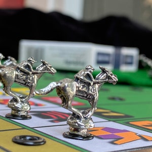 May include: Silver horse racing game pieces on a green felt game board. The horses are in different positions on the board, with one horse in the lead. The board has black circles and a yellow and purple jockey silks.