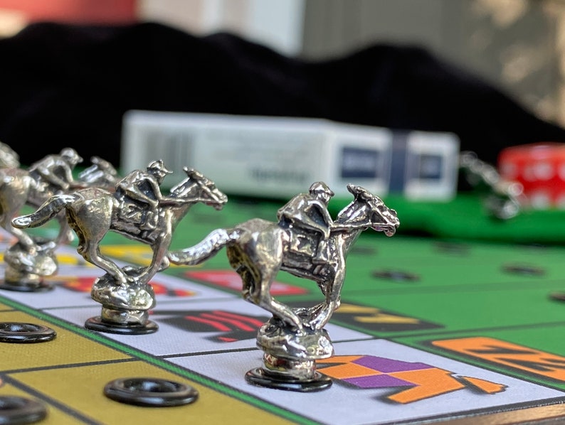 May include: Silver horse racing game pieces on a green felt game board. The horses are in different positions on the board, with one horse in the lead. The board has black circles and a yellow and purple jockey silks.