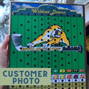 May include: A green and yellow Wildcat Downs game board with a yellow wildcat mascot holding a hockey stick. The board has a grid of holes and numbers 8 through 12 at the bottom.